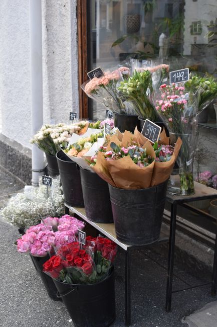 Display of various fresh flower bouquets outside a flower shop, arranged in black buckets on a two-tier black metal stand. The flowers include pink roses, white carnations, red roses, and mixed pink a