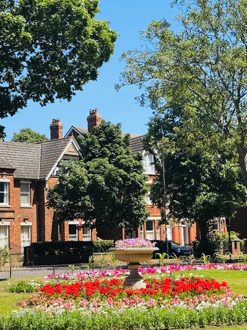 A vibrant public space featuring a large, ornate stone planter filled with pink and purple flowers, surrounded by well-maintained flowerbeds with red, pink, and white blooms. In the background, tradit