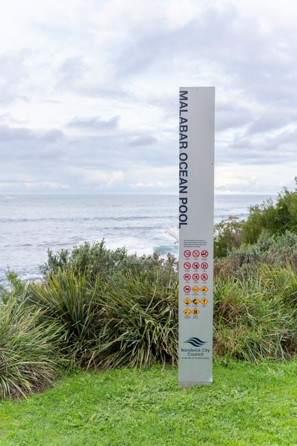A tall, slim, rectangular signpost with a white background and black text reading 'Malabar Ocean Pool' standing on a grassy, landscaped area near the coast. Surrounding the sign are green, spiky coast
