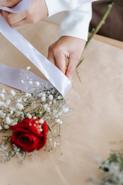 A person’s hands are seen wrapping a bridal bouquet featuring a vibrant red rose surrounded by delicate white baby's breath and sprigs of greenery. The bouquet is being carefully arranged on a light